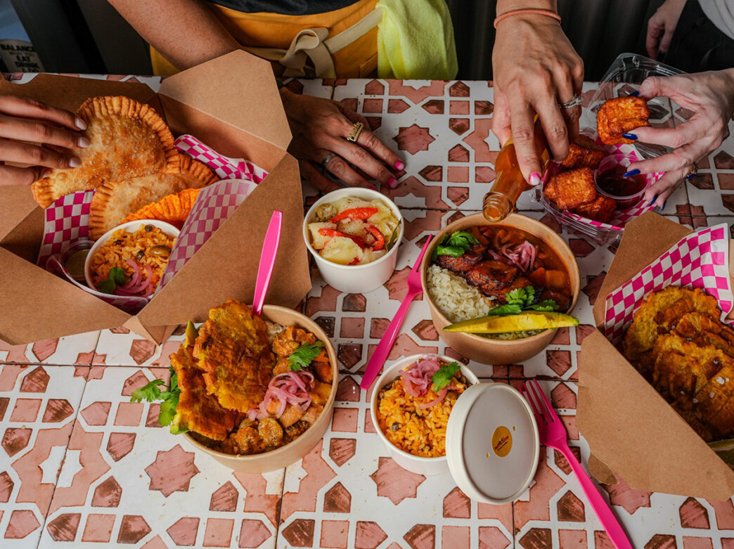 Pastelillos, rice and other meals are displayed on a pink and white tabletop. People's hands reach for food and pour hot sauce over the food.