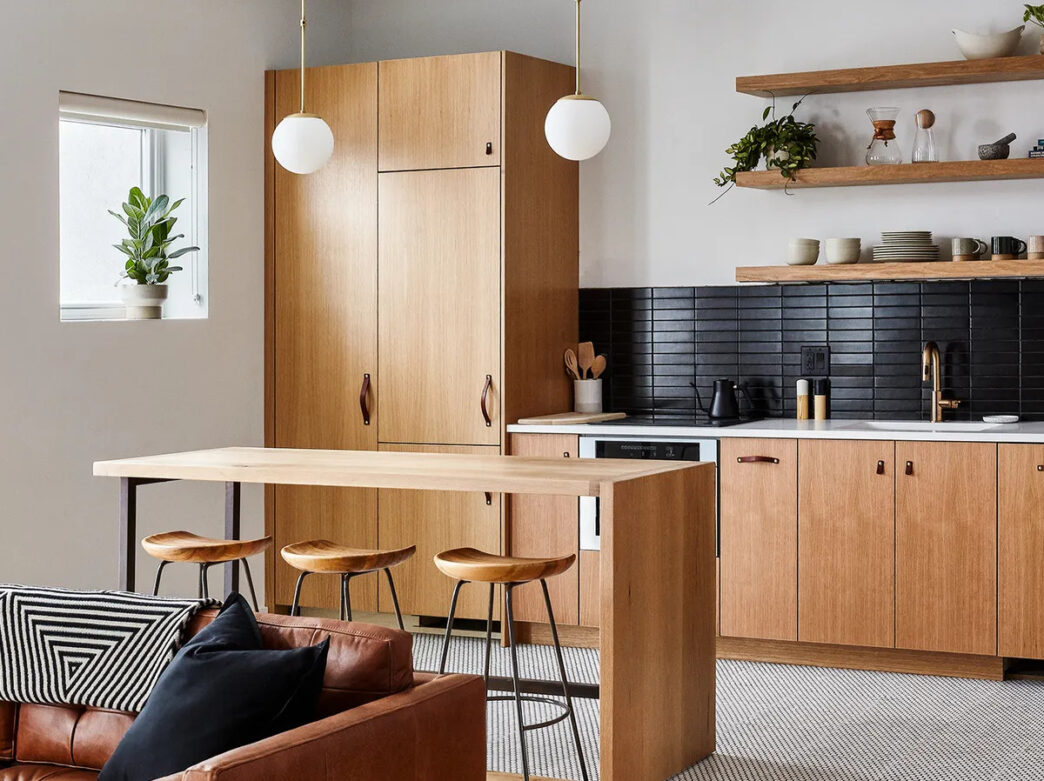 A kitchen with natural wood cabinets, black tile backslap and open shelving at Lokal Hotel Fishtown. The room also features a wood island with 3 barstools and a leather couch.