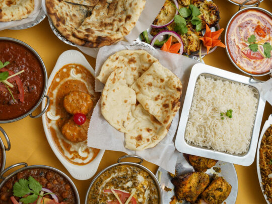 Rice, naan and a variety of Indian dishes are displayed on a table with a yellow table cloth.