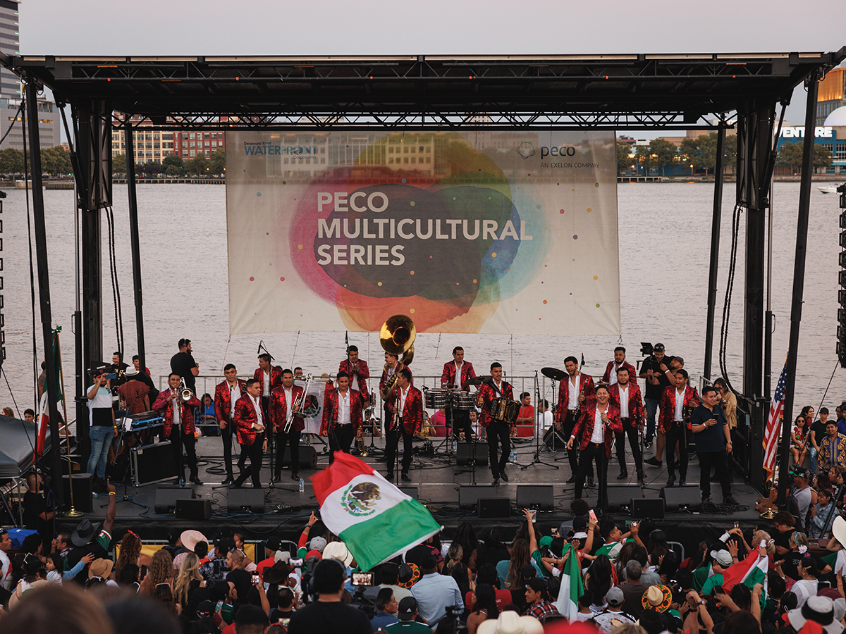 Un groupe joue sur la scène du festival de la fête de l'indépendance mexicaine, devant le fleuve Delaware, tandis que les festivaliers dansent et écoutent de la musique.
