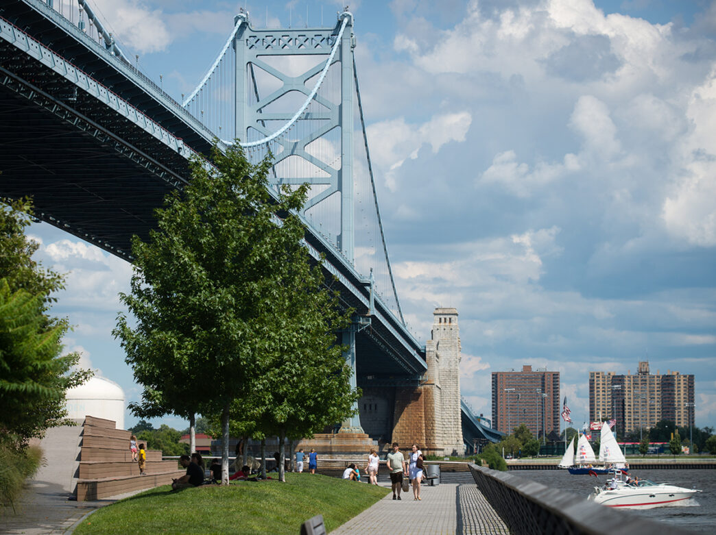 People lounge and walk along the Race Street Pier overlooking the Delaware River and Benjamin Franklin Bridge on a sunny day.