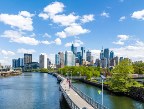 The Philadelphia skyline shines under a blue sky with white clouds. IN the foreground, the Schuylkill Banks Boardwalk juts out over the Schuylkill River