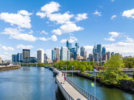 The Philadelphia skyline shines under a blue sky with white clouds. IN the foreground, the Schuylkill Banks Boardwalk juts out over the Schuylkill River