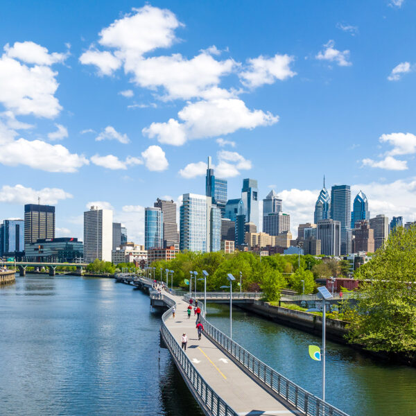 The Philadelphia skyline shines under a blue sky with white clouds. IN the foreground, the Schuylkill Banks Boardwalk juts out over the Schuylkill River