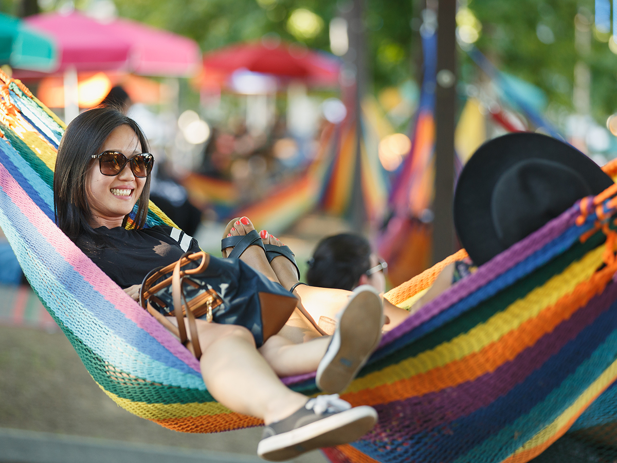 Two people lay head-to-toe in a colorful rainbow hammock at Spruce Street Harbor Park during the day.