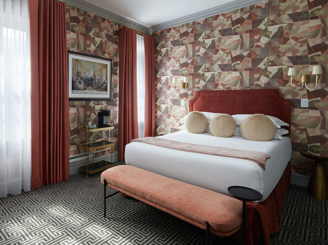 A queen guest room with white linens with a rust red velvet headboard. The room has a geometric green and rust red wallpaper and floor to ceiling curtains.