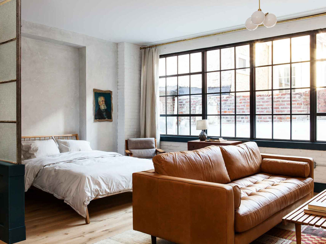 A guest room loft at the Lokal Old City with a midcentury style bed and brown leather couch. The room has wall to wall windows and hanging globe light fixture hanging from the ceiling.