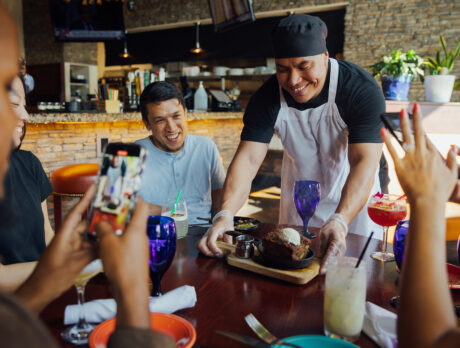 The chef at El Sarape, wearing a black chef's hat and apron, delivers a dessert to a table of diners who are capturing the moment using their phones.