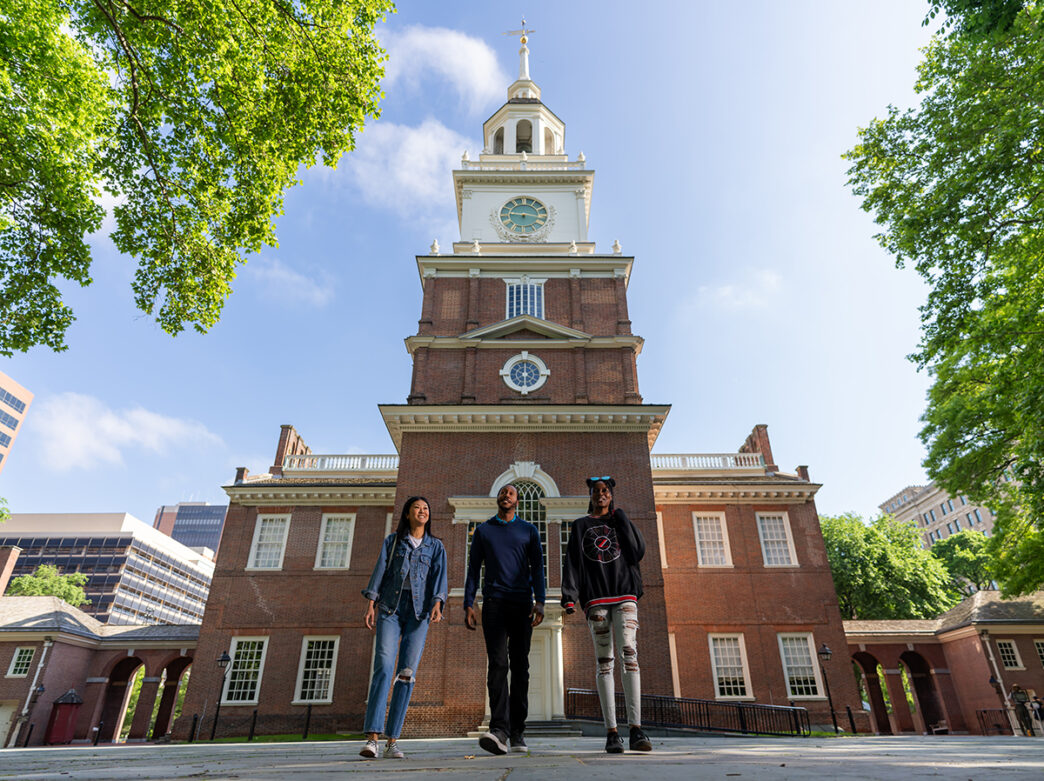 Three people walk with the back of Philadelphia's Independence Hall in the background.