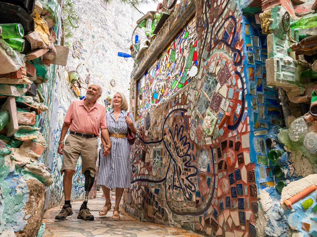 Un homme portant un short et un polo rose tient la main d'une femme vêtue d'une robe rayée et de sandales. Ensemble, ils traversent les murs en mosaïque des Magic Gardens de Philadelphie.