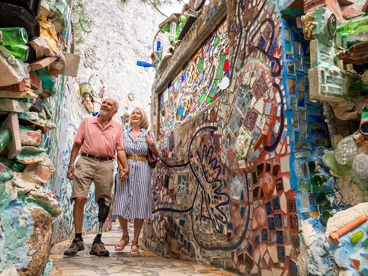 A man wearing shorts and pink polo shirt holds the hand of a woman wearing a stripped dress and sandals. Together, they walk through the mosaic walls at Philadelphia's Magic Gardens.
