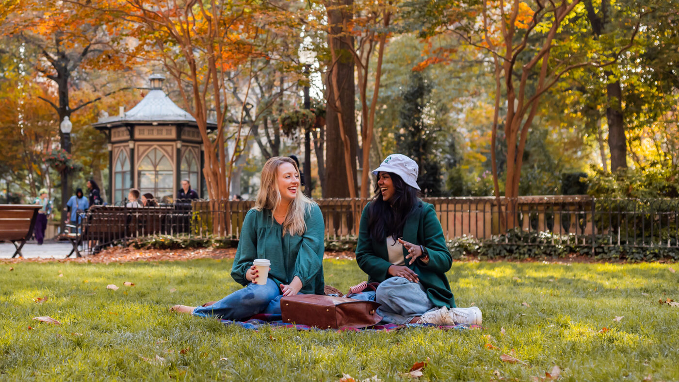 Two women sit in the grass together drinking coffee and laughing in Rittenhouse Square on a fall day.