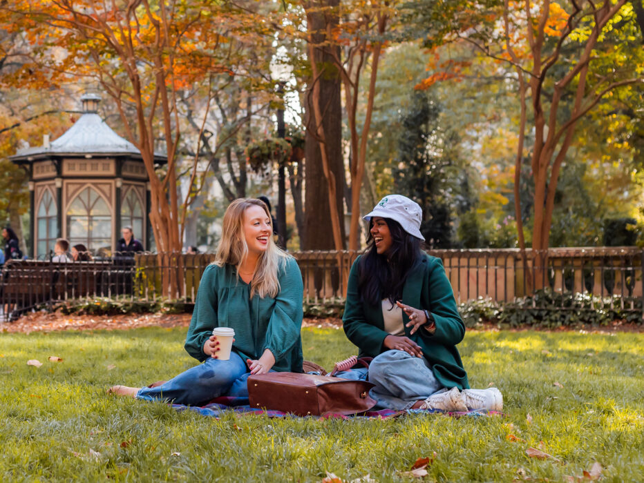 Two women sit in the grass together drinking coffee and laughing in Rittenhouse Square on a fall day.