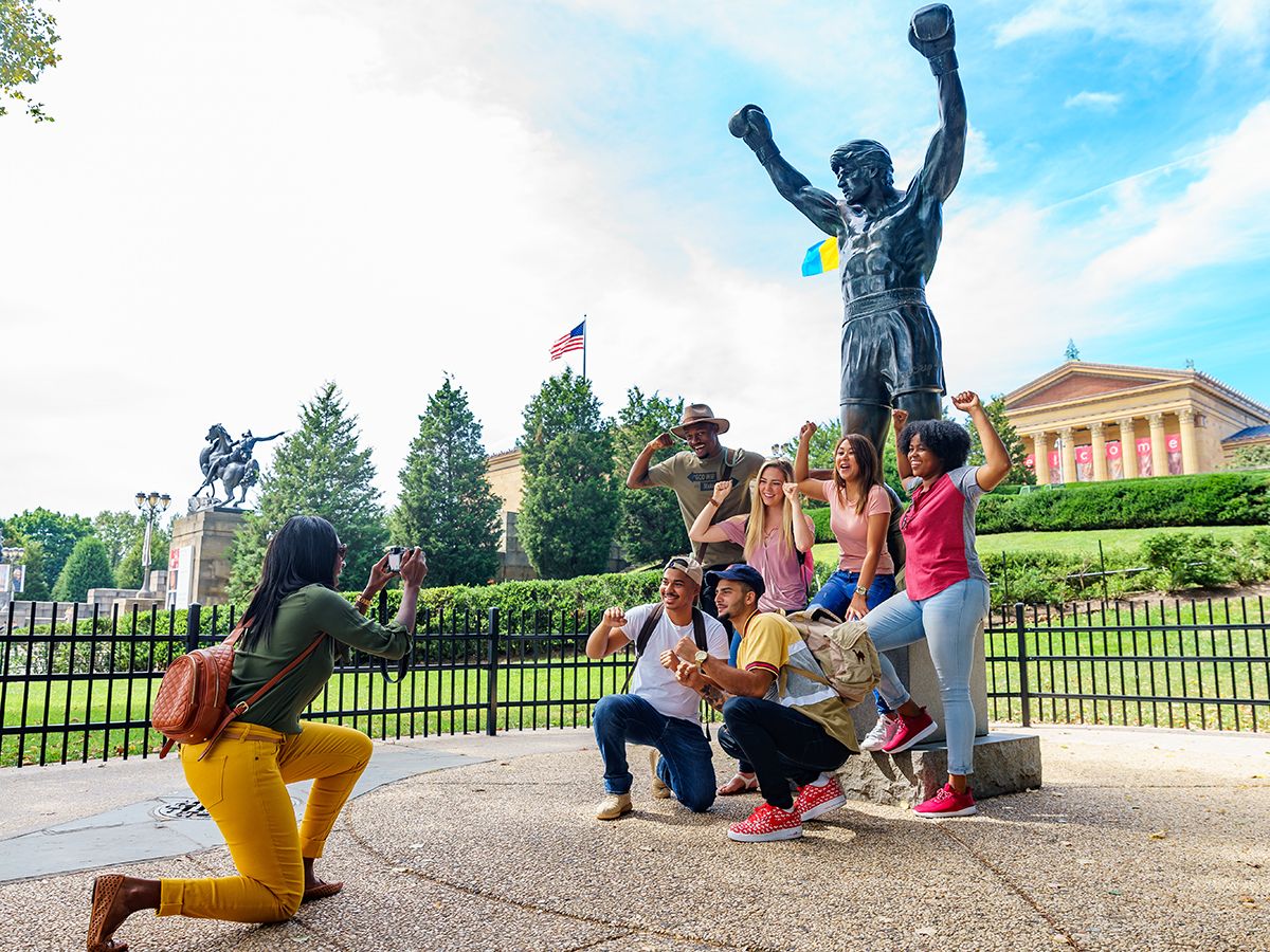 A group of six people pose for a photo in front of a bronze statue of Rocky at the Philadelphia Museum of Art.