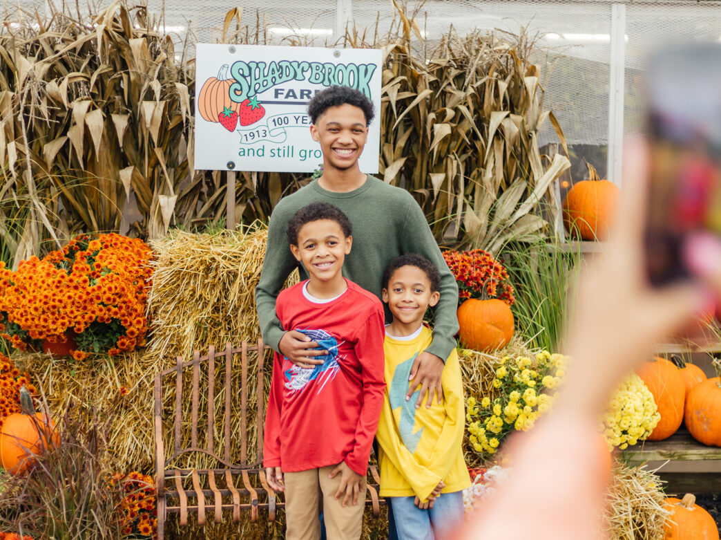 Three kids pose together in front of stacks of hay, mums, cornstalks and pumpkins at Shady Brook Farm while someone takes their photo with a phone.