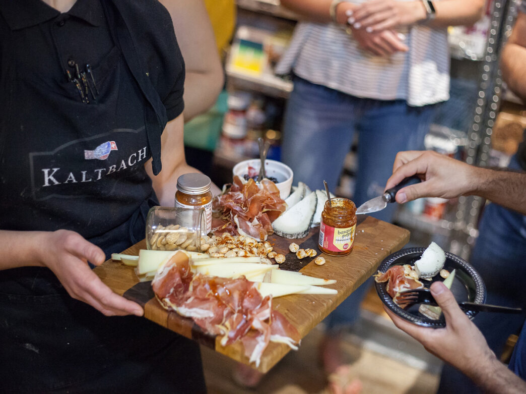 Una persona con un delantal negro sostiene una tabla de embutidos con carne, queso, frutos secos y mermelada, mientras otra persona selecciona los productos de la tabla.