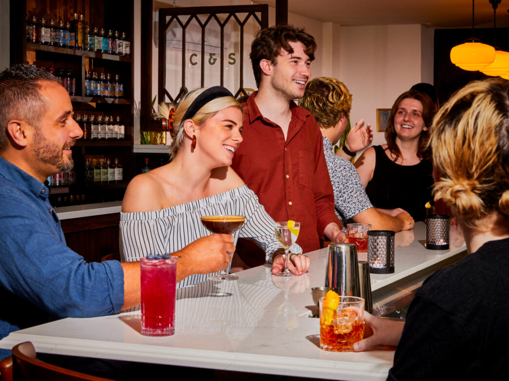 La gente se sienta en una barra con una superficie de mármol blanco mientras sonríe y sostiene cócteles en Char and Stave.