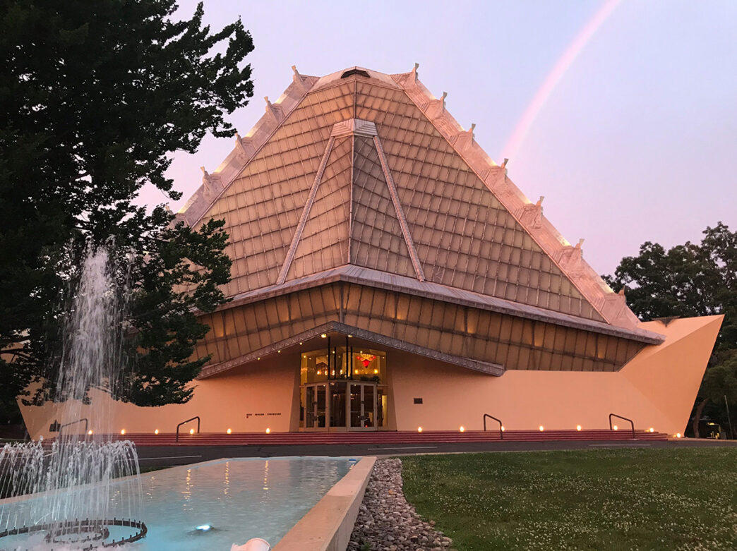 An exterior view of Beth Sholom Synagogue with a decorative fountain in the foreground and a rainbow in the sky.