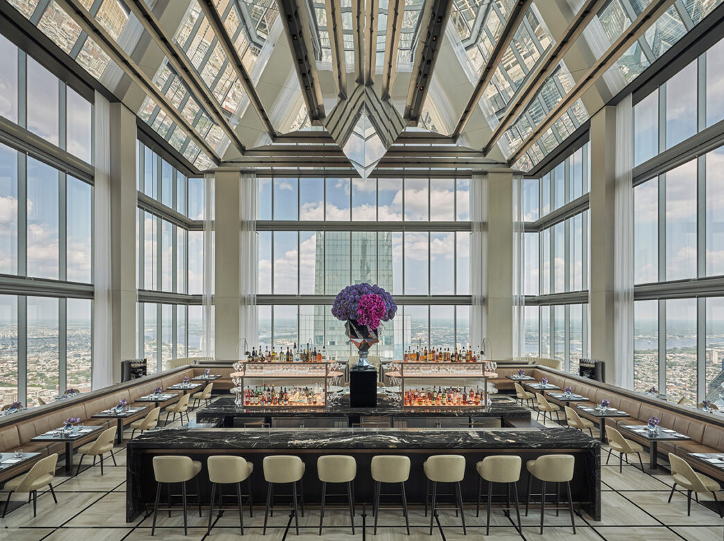 A rooftop restaurant and bar with a glass ceiling and floor to ceiling windows. Tables and chairs surround a marble bar.