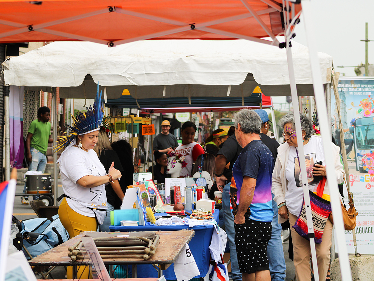 A vendor wearing a feather headdress stands behind a table while festival attendees look at items displayed on the table.