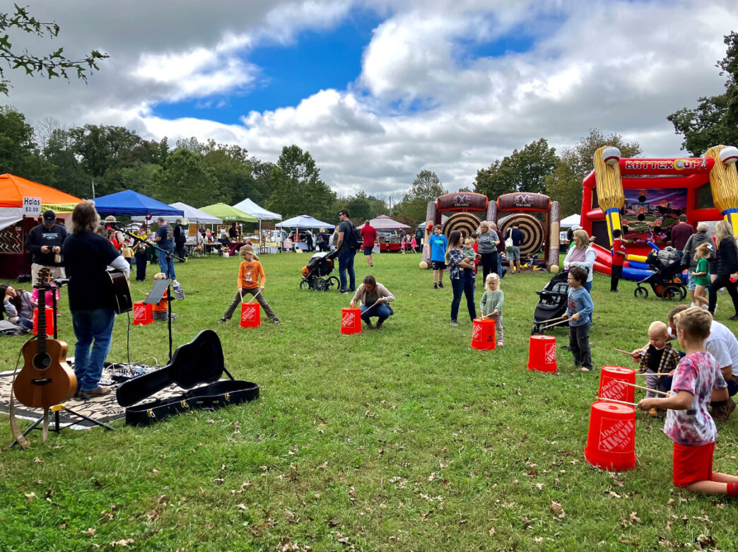 Kids use drumsticks on buckets to play along with a guitarist at the Warwick Fall Festival. Inflatable games and vendor tents are seen in the background.