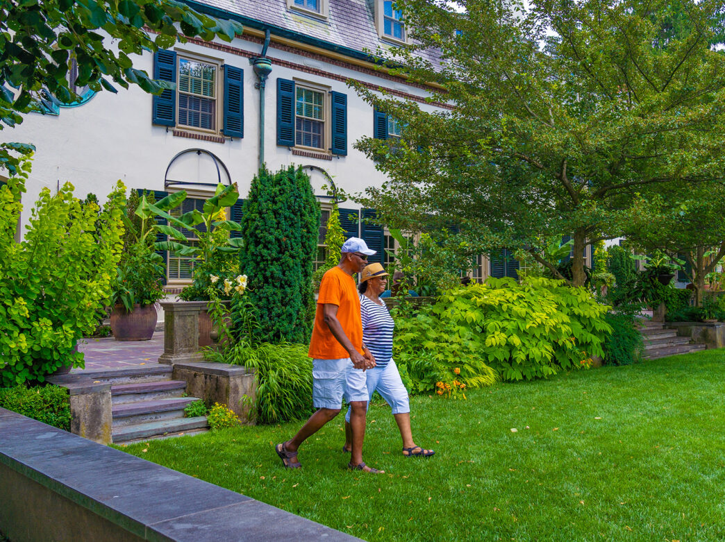 Two people wearing shorts, hats and sandals walks through a grass lawn in front of a historical building at Chanticleer.