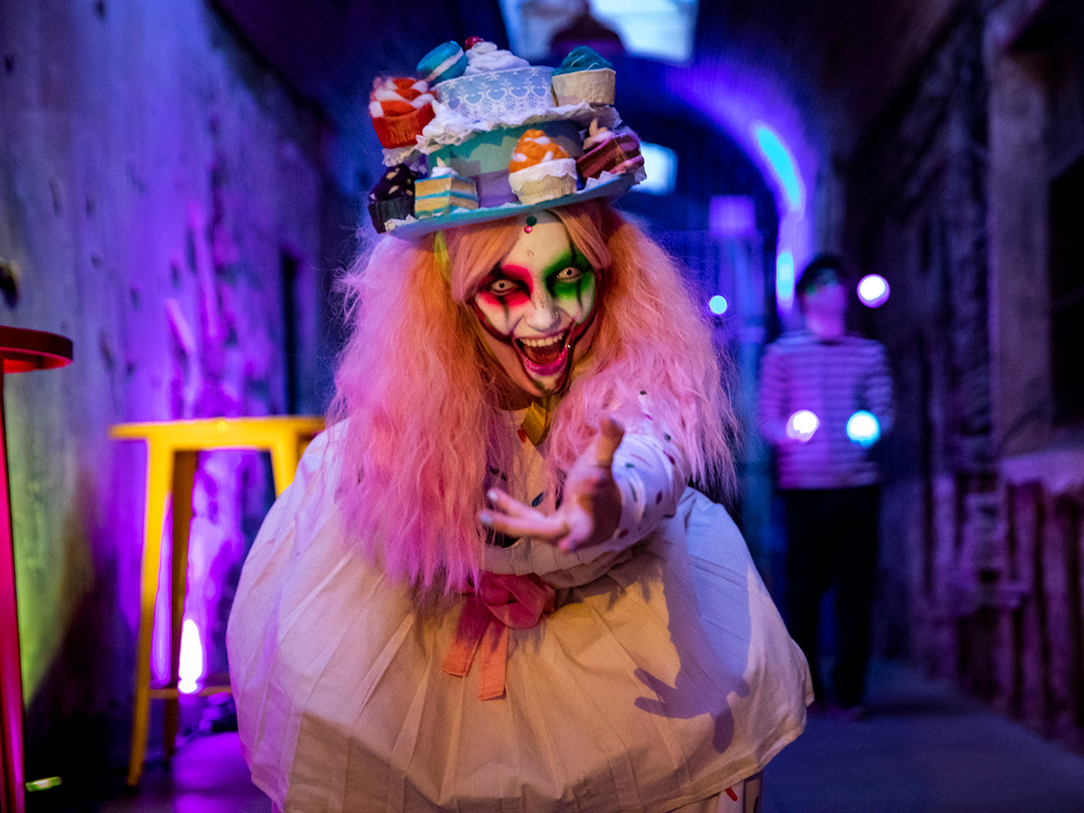 A creepy clown, wearing a pastel pink wig topped with a hat full of devilishly delicious-looking desserts, smiles and beckons you into the haunted house at Eastern State Penitentiary.