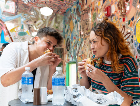 Two people stand at a table and eat cheesesteaks at Jim's Steaks on South Street. Behind them are colorful mosaics.