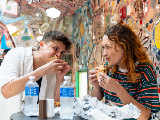 Two people stand at a table and eat cheesesteaks at Jim's Steaks on South Street. Behind them are colorful mosaics.