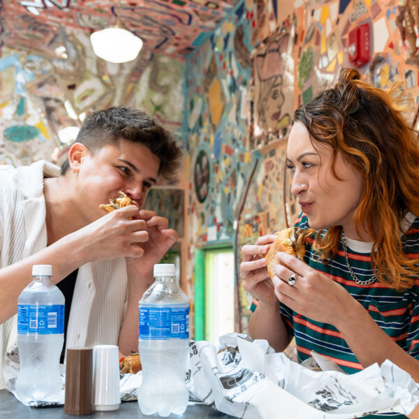 Two people stand at a table and eat cheesesteaks at Jim's Steaks on South Street. Behind them are colorful mosaics.