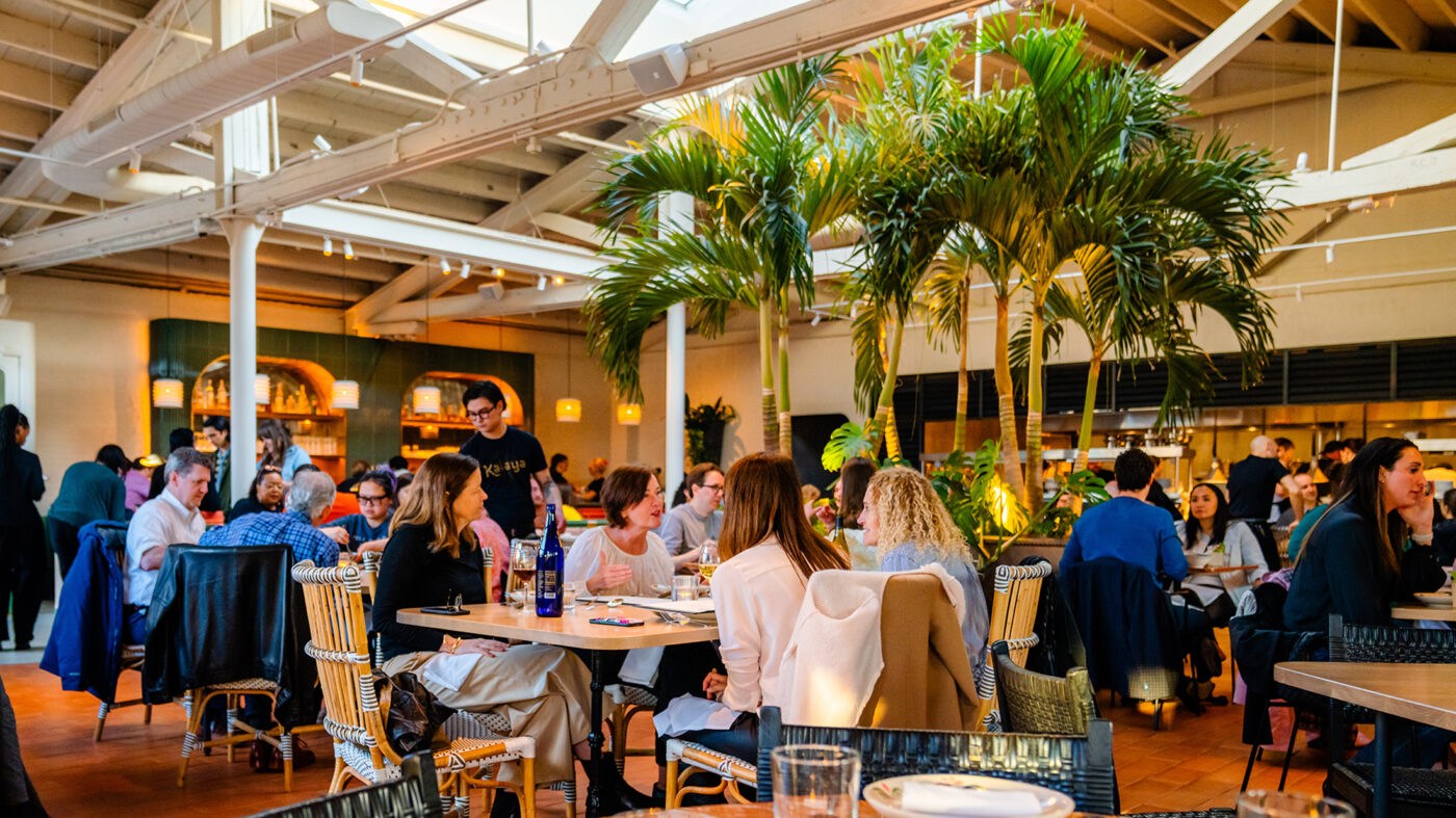 Les convives s'assoient à des tables et dégustent des plats et des boissons dans la salle à manger de Kalaya. Des palmiers aussi hauts que le plafond se trouvent au centre de la salle à manger.