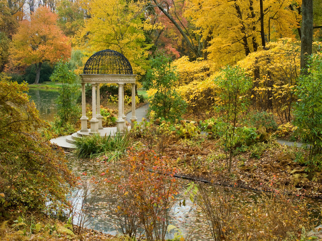 Trees with yellow and orange leaves surround a stone and iron gazebo at the edge of a lake at Longwood Gardens.