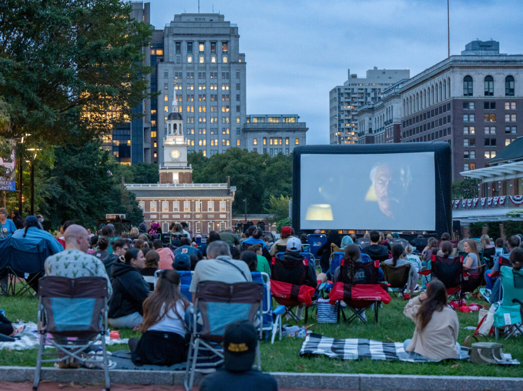 People sit in camping chairs and on blankets in the grass on Independence Mall for Movies on the Mall. A large screen displays a movie. Independence Hall can be seen in the background.
