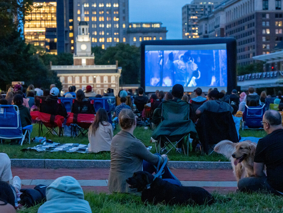 La gente se sienta en sillas de camping y sobre mantas con perros en el césped del Independence Mall para ver Movies on the Mall. Una gran pantalla proyecta una película. Independence Hall se ve al fondo.