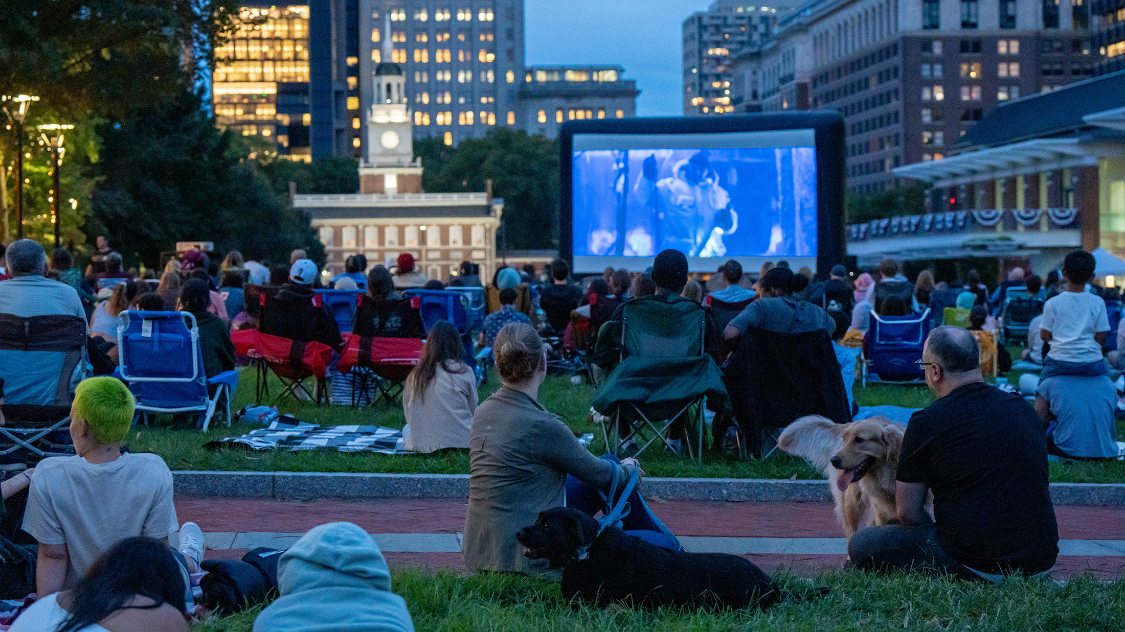 People sit in camping chairs and on blankets with dogs in the grass on Independence Mall for Movies on the Mall. A large screen displays a movie. Independence Hall can be seen in the background.