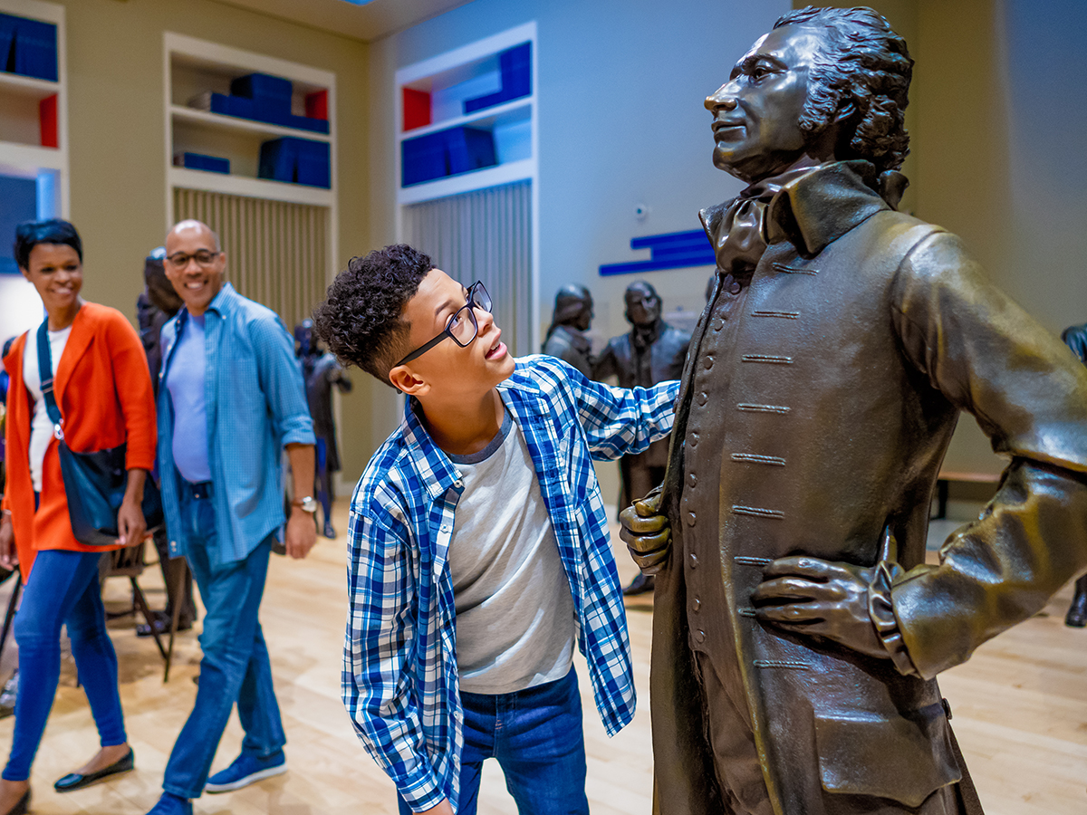 A kid wearing a blue and white checkered shirt looks up at a bronze statue of Alexander Hamilton in the Signer's Hall exhibit at the National Constitution Center.