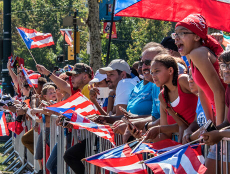 People watching the Puerto Rican Day Parade wave handheld Puerto Rican flags while standing behind a metal fence.
