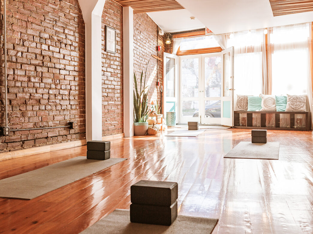 The interior of a yoga studio with hardwood floors and exposed brick walls. Yoga mats with yoga blocks are laid out on the floor.