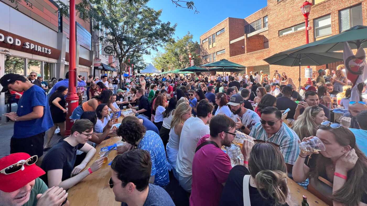 People sit at long wood picnic tables in the street while drinking beer and talking with friends at the annual South Street Oktoberfest.