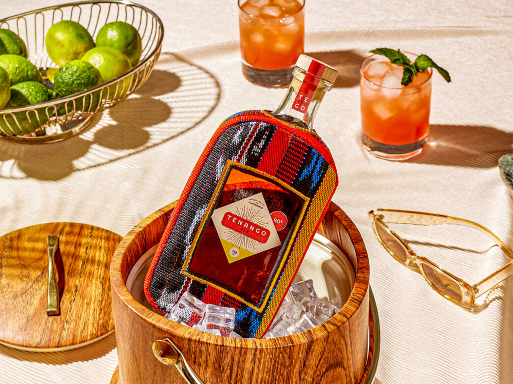 A bottle of rum is displayed in an ice bucket. Cocktails. a basket of limes and a pair of sunglasses are in the background.