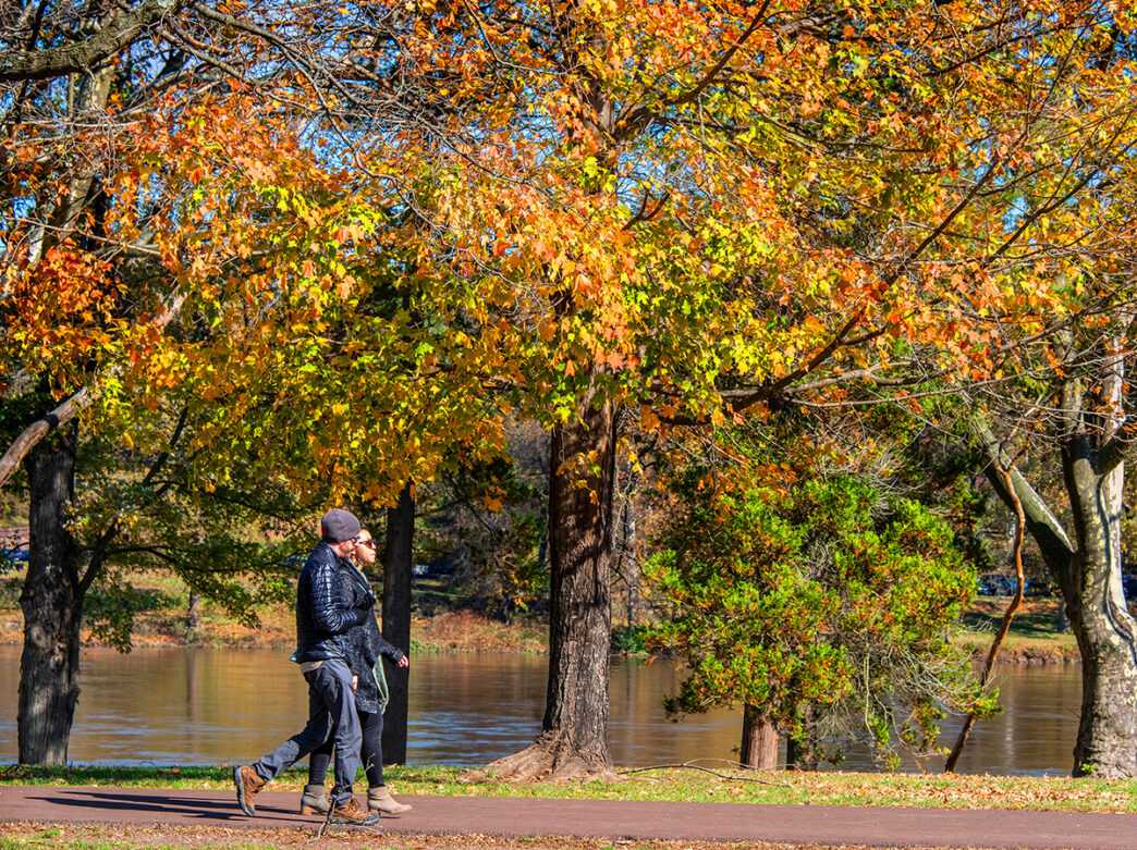 Two people wearing jackets and boots walk along a paved path adjacent to the Delaware River. Trees with yellow, orange and red leaves line the path.