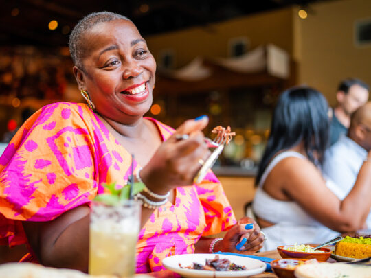 A person wearing a pink and orange dress holds up a forkful of meat while dining at Zahav.