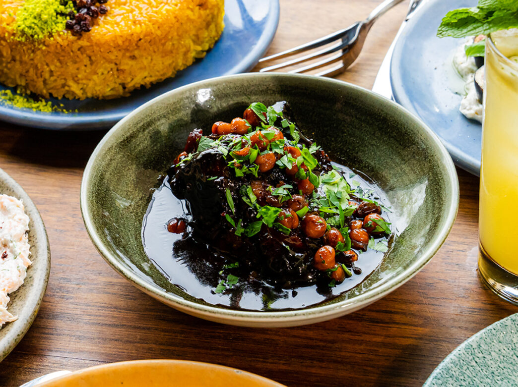 Lamb shoulder glazed with pomegranate molasses and topped with chickpeas is displayed in a sage green dish on a tabletop with other menu items at Zahav.
