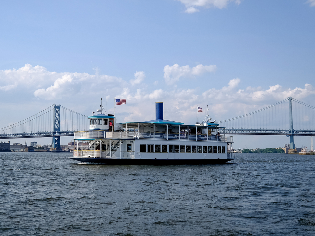 A white and blue ferry travels across the Delaware River with the Benjamin Franklin Bridge in the background.