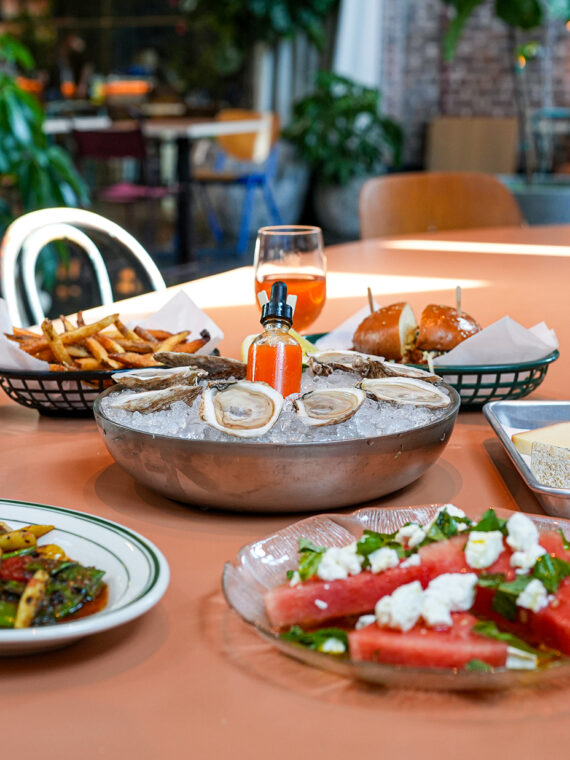 A variety of menu items are displayed on a salmon tabletop with chairs and floor pants in the background. Menu items include oysters, watermelon salad, chicken sandwich, fries, cocktails and glasses of wine.