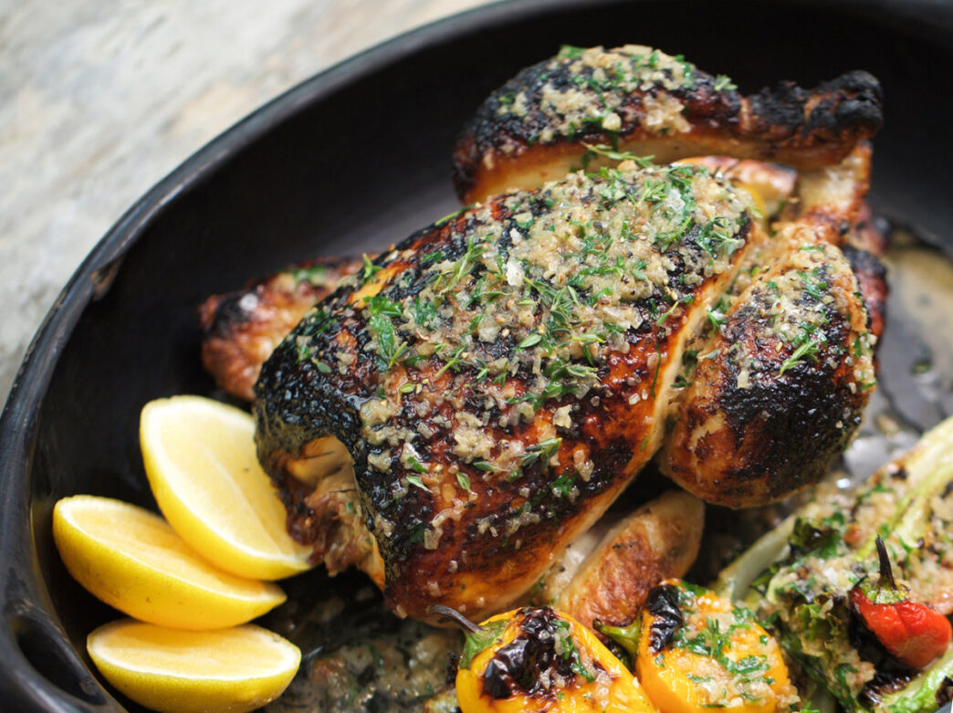 A whole roast chicken topped with herbs and lemon is served in a cast iron dish with roasted vegetables and lemon slices.