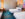 A bright and stylish hotel room featuring two beds with white linens, a colorful accent pillow, a wooden headboard with integrated lighting, and a vibrant blue carpet. A small workspace with a desk and orange chair is tucked into the corner.