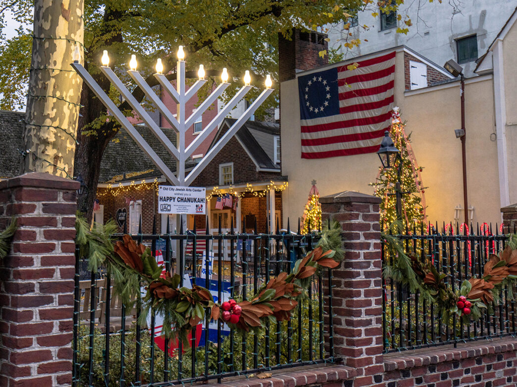 A menorah is lit outside of the Betsy Ross House in Philadelphia at dusk. Behind, a historic American flag hangs on a building and holiday lights are draped on the building.