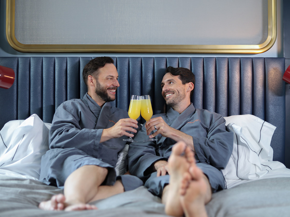 A smiling couple lays in bed and toasts their mimosas while wearing matching gray linen robes.
