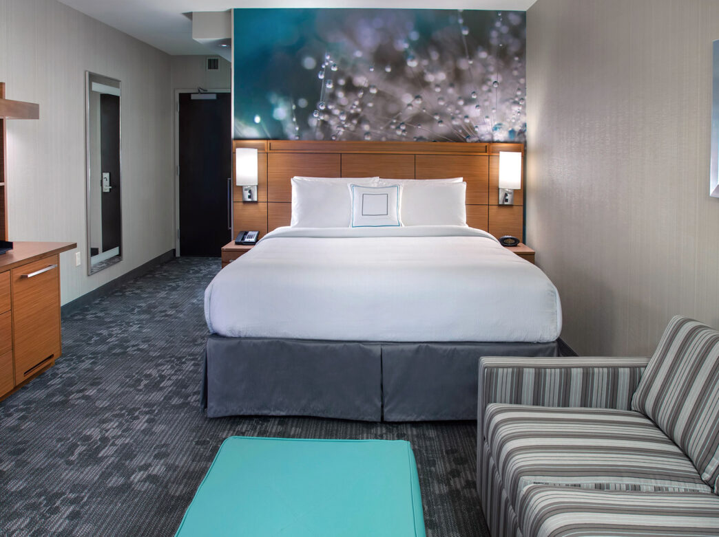 Une chambre d'hôtel à l'intérieur du Courtyard South Philadelphia at the Navy Yard avec un lit king-size blanc et gris, un canapé rayé, une petite table bleu sarcelle et une télévision sur une commode.